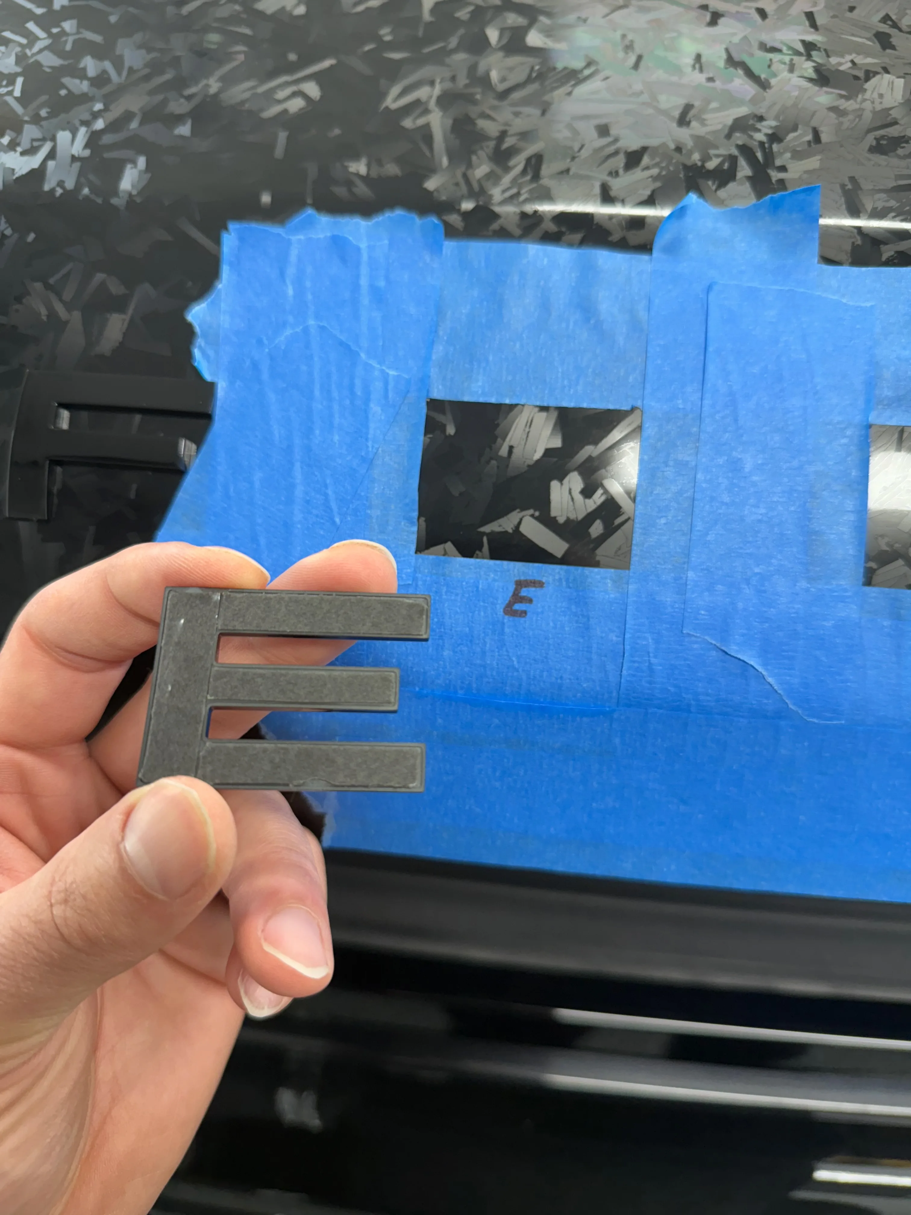Vehicle emblem removed with blue masking tape revealing PPF installed underneath — attention to detail during paint protection film installation